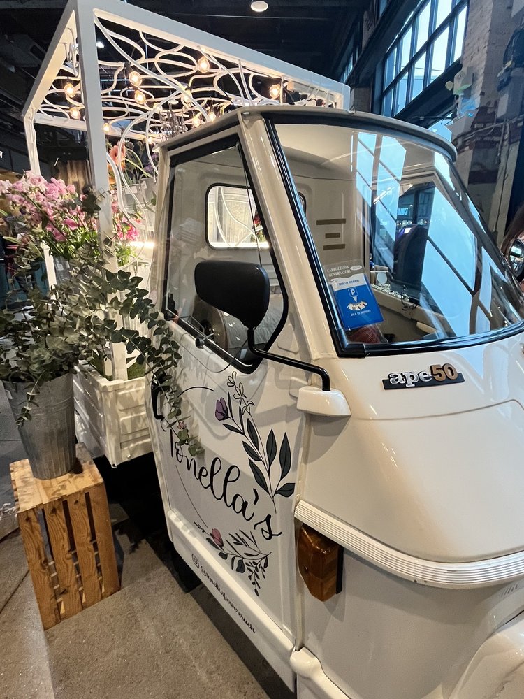 Tonella's vintage white Piaggio Ape flower truck, cursive script and botanical line-art on the door, fresh blooms in a zinc bucket beside it